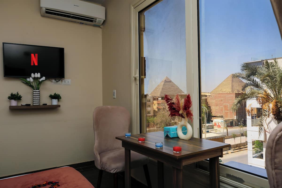 A hotel room with a flat-screen TV mounted on the wall, a window with a view of pyramids, a table with candles, and a chair.
