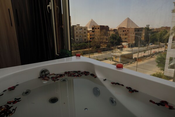 A bathroom with a bathtub filled with rose petals, a window with curtains, and a view of the pyramids in the background. - Hammis Pyramids View  (Giza)