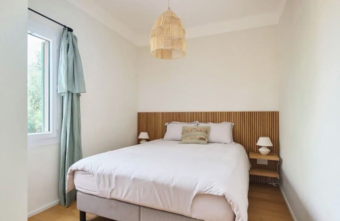 Un dormitorio con cama, lámparas de noche, una lámpara colgante, una ventana con cortinas y vistas al exterior.