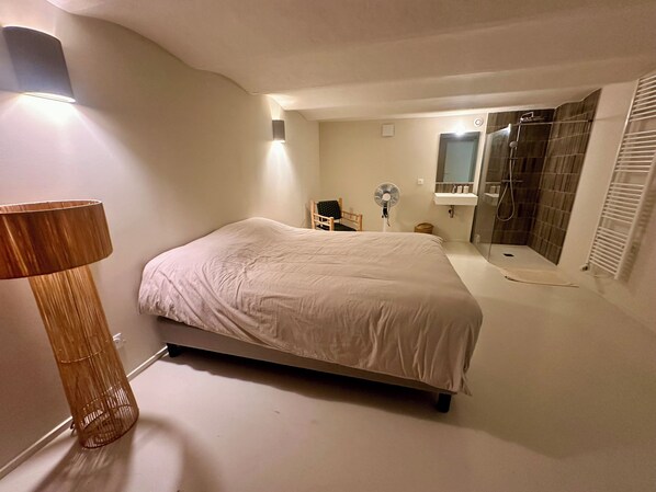 A bedroom with a bed, a bedside table, a lamp, a fan, a chair, a mirror, and a shower area.