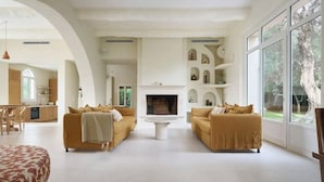 A spacious living room with two sofas facing a fireplace, a dining area with a table and chairs, and large windows offering a view of the outdoors. - Villa El Sol Juan Les Pins (Antibes)