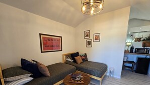 A cozy living room with a sofa, a coffee table, and a checkered rug. There is a framed picture on the wall and a kitchen area in the background.