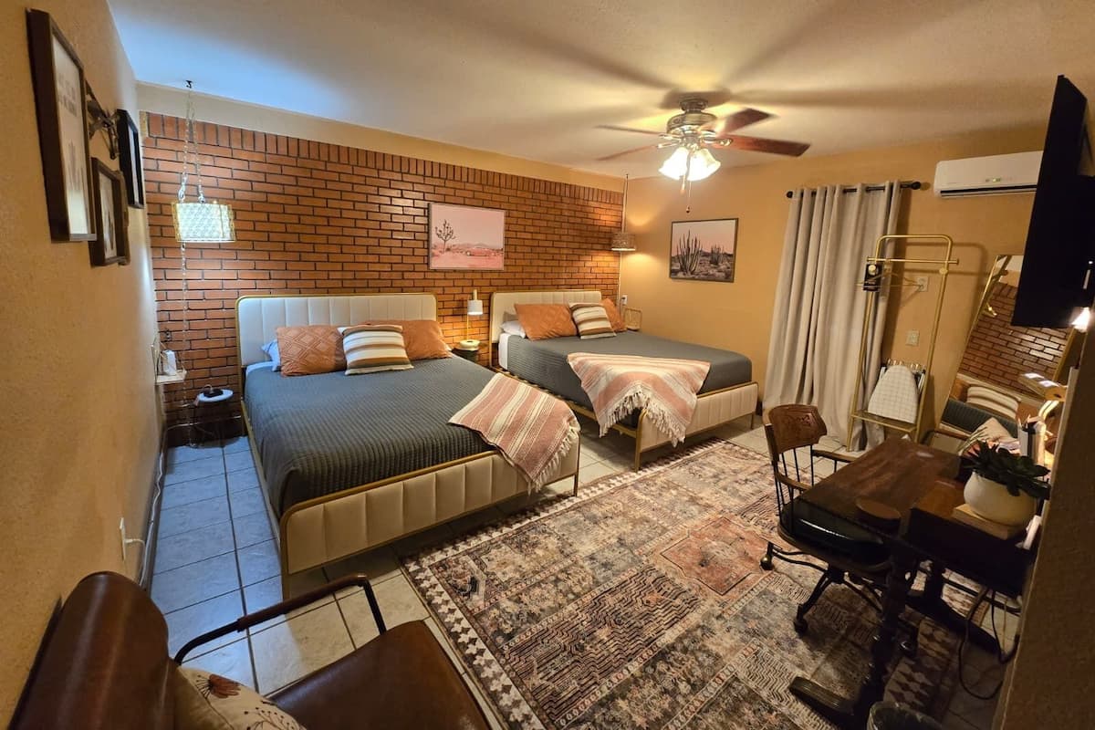A hotel room with two beds, a desk, a chair, a ceiling fan, and a brick wall.