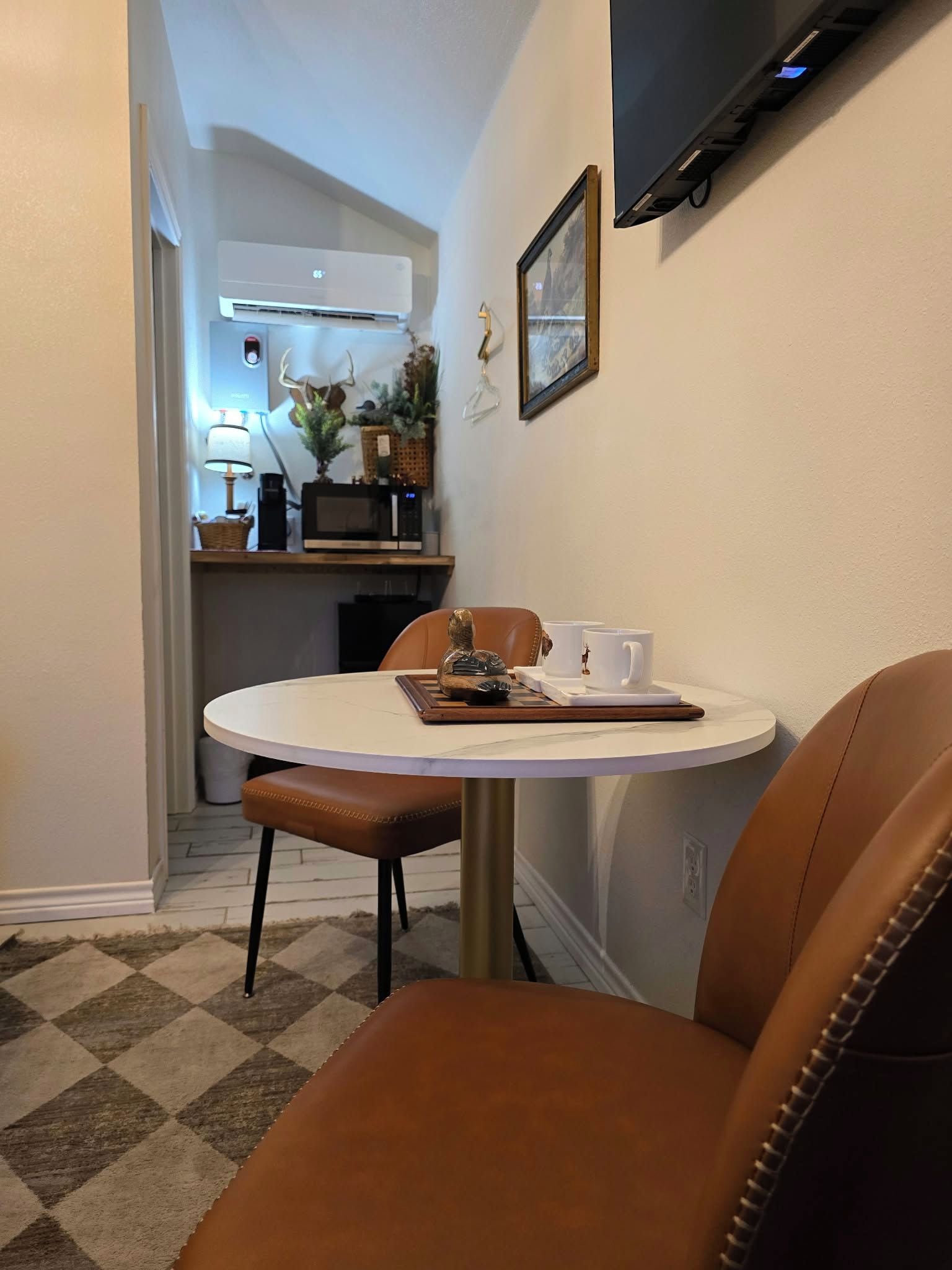 Un comedor con una mesa redonda, dos sillas, una bandeja con un objeto decorativo y un televisor montado en la pared.