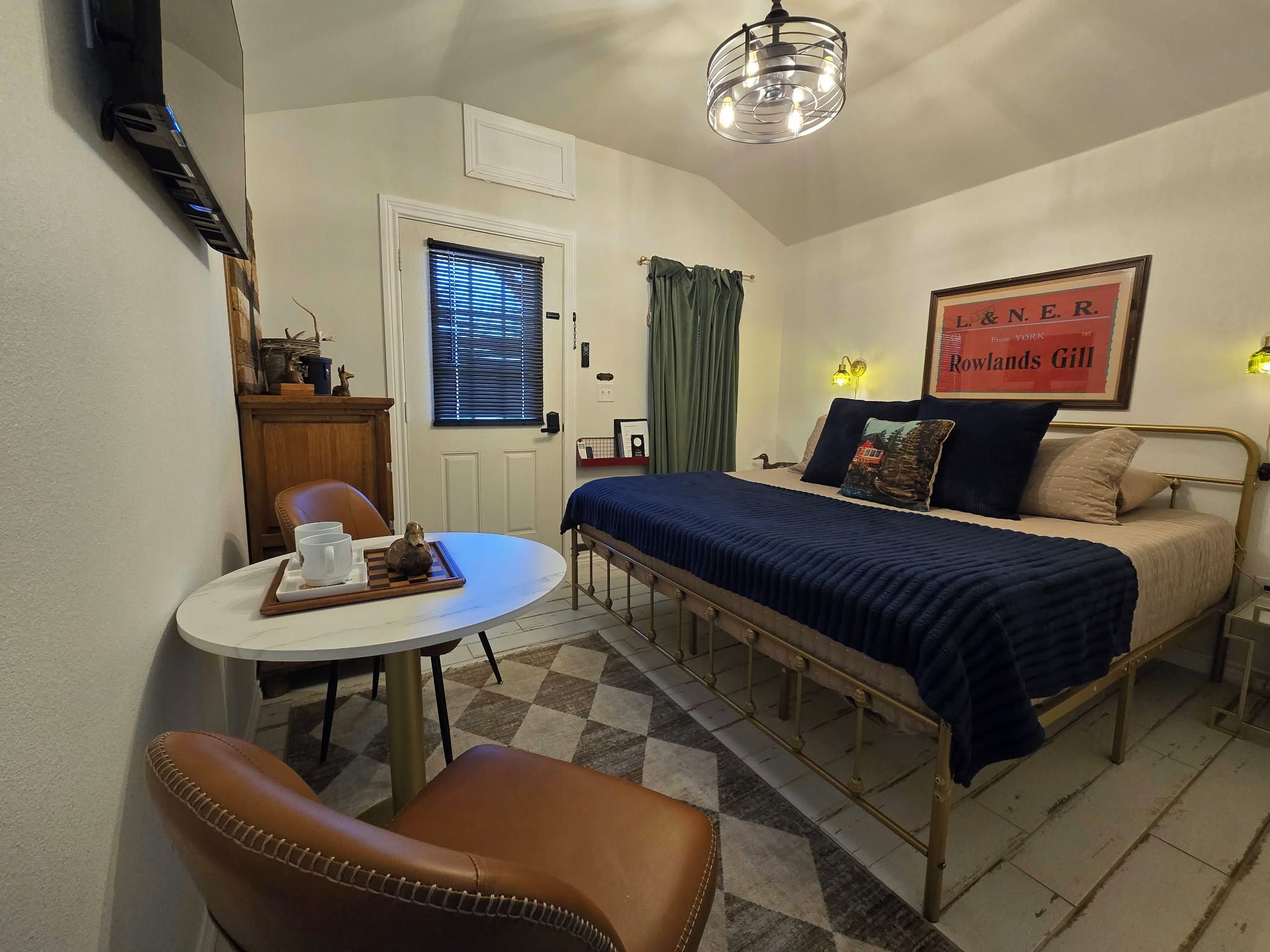 Une pièce de taille modeste comprenant un lit, une petite table avec des tasses et un plateau, une chaise, un bureau avec un ordinateur, une fenêtre avec des stores, un rideau, une lampe fixée au mur et un panneau décoratif encadré.