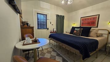 A compact room with a bed, a small table with cups and a tray, a chair, a desk with a computer, a window with blinds, a curtain, a wall-mounted light, and a framed sign.