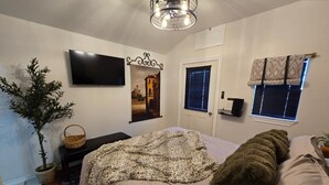 A bedroom with a bed, a TV, a wall-mounted picture, a door, and a window with blinds.