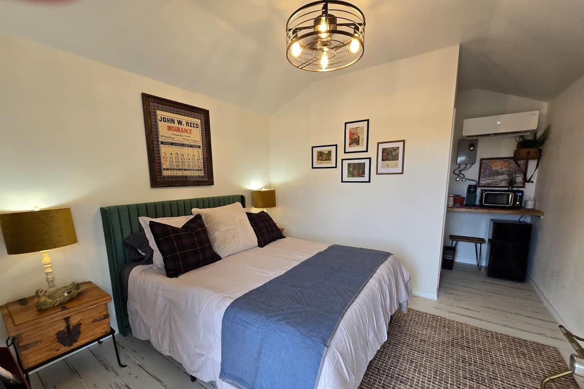 A neatly arranged bedroom with a bed, bedside lamps, a framed poster, and a small kitchenette area.