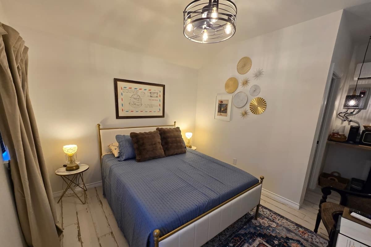 A bedroom with a bed, a small bedside table, a lamp, a wall-mounted picture, and a ceiling light fixture.