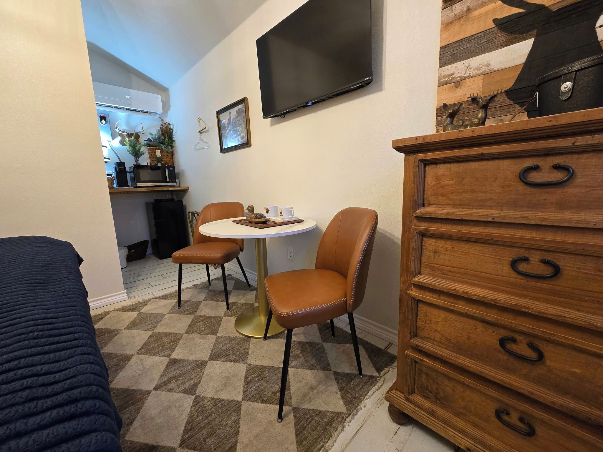 Een gezellig, compacte leefruimte met een eethoek, een aan de muur bevestigde televisie en een houten dressoir met lades.