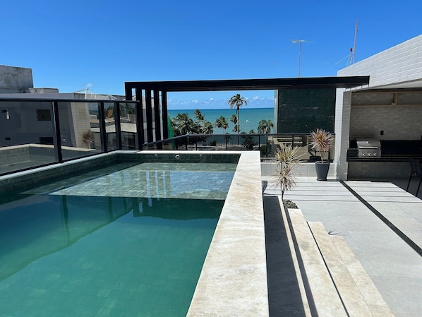 An outdoor swimming pool with a view of the ocean, a barbecue area, and a potted plant. - Bela Hospedagem - Mahalo Flats a 80m da praia (João Pessoa)