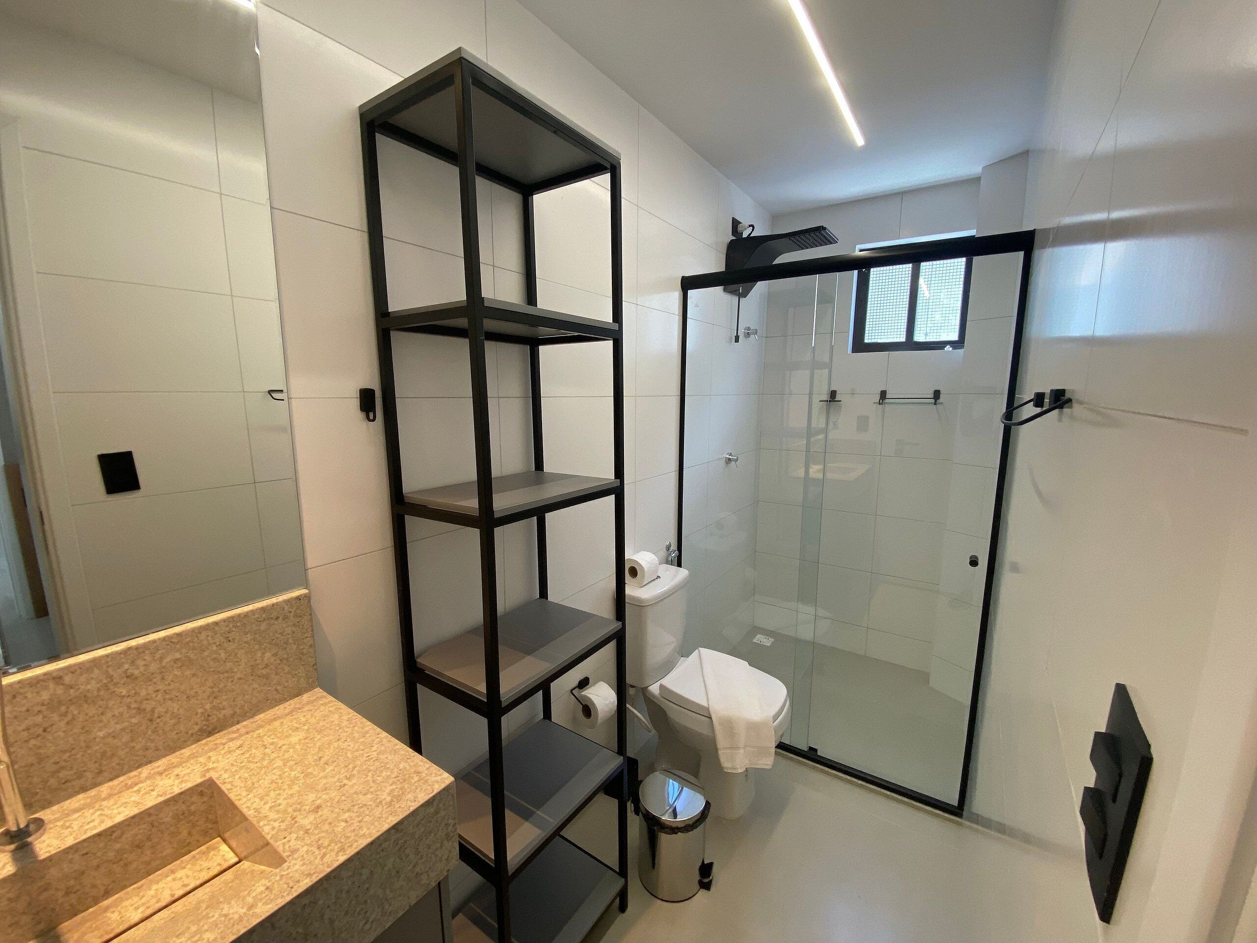 Ein Badezimmer mit Duschkabine aus Glas, Toilette, Waschbecken mit Arbeitsplatte und einem Metallregal.