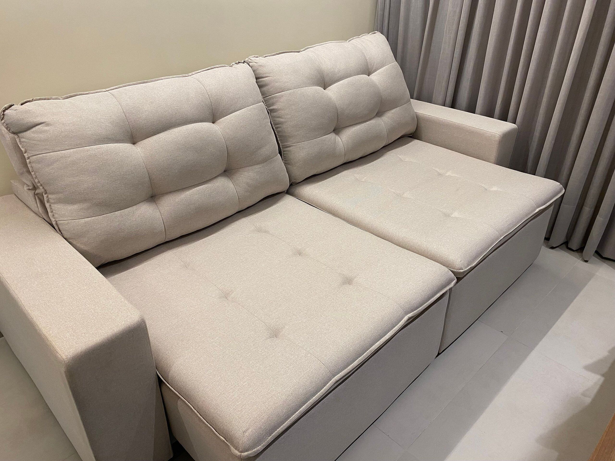 A beige sofa with a cushioned backrest and armrests, positioned against a wall with a window and curtains.