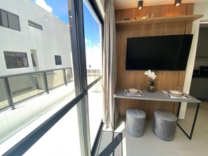 A modern interior with a large window, a flat-screen TV, a small table with a vase, and two stools. - Bela Hospedagem - Mahalo Flats a 80m da praia (João Pessoa)