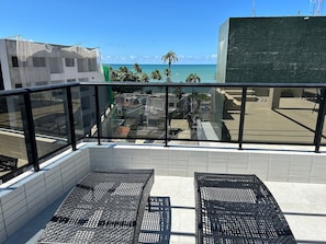 A balcony with two wicker chairs, a view of the ocean, and a building with a tiled facade. - Bela Hospedagem - Mahalo Flats a 80m da praia (João Pessoa)