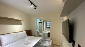 Quarto de hotel com cama, televisão, espelho e área de chuveiro.