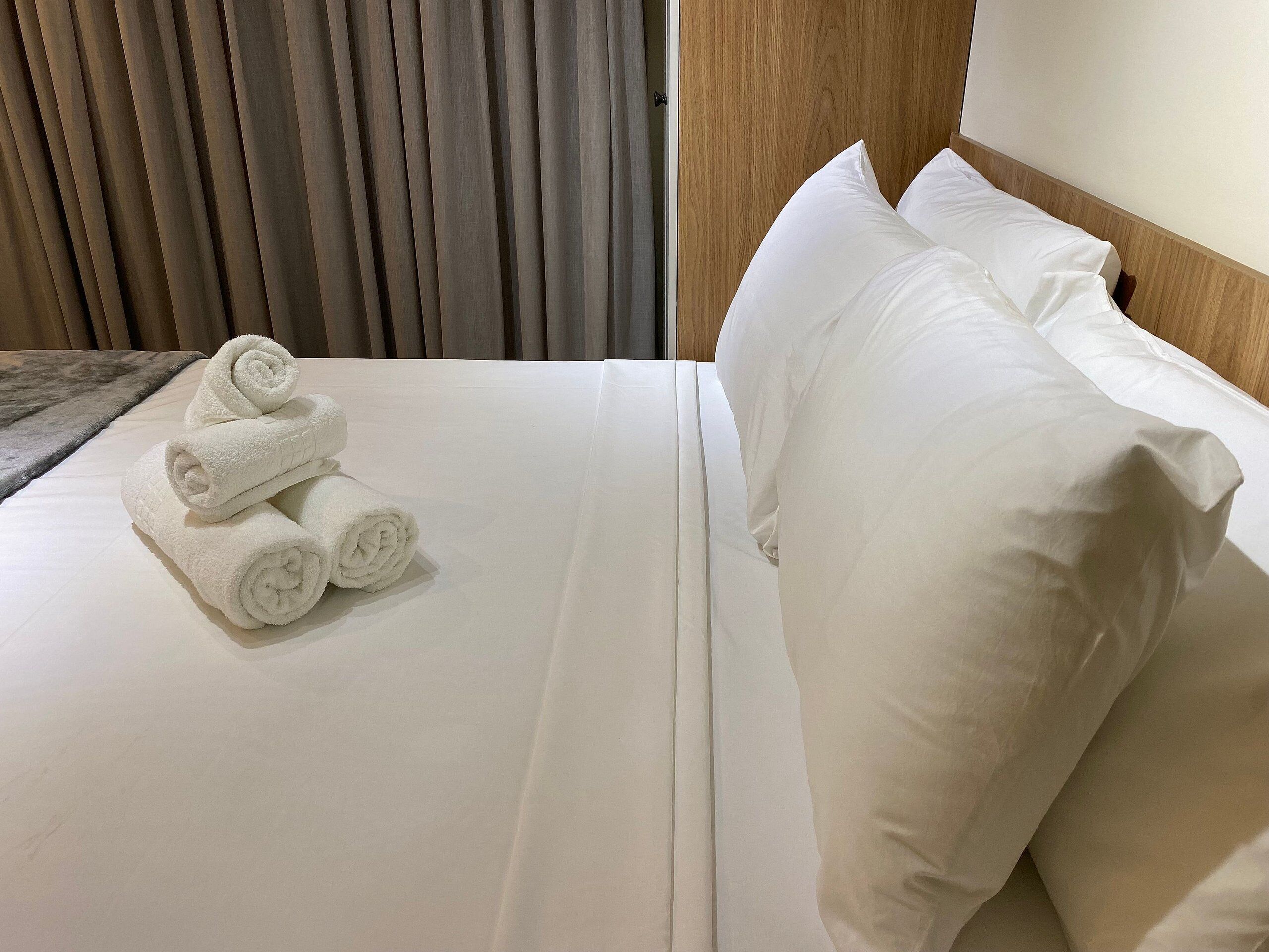 A neatly made bed with white linens and pillows, a rolled white towel on the bed, and a wooden headboard.