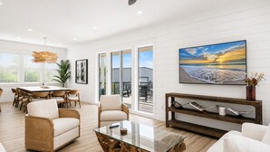 A spacious living room with a glass coffee table, wicker furniture, a large painting of a beach, and a view to the outside. - Casa Blanca | Rooftop Bar • Elevator • Golf Cart • 400' Beach Access (Miramar Beach)
