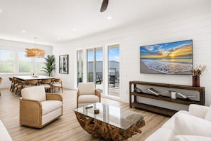 Amplia sala de estar con una mesa de centro de vidrio, muebles de mimbre, un cuadro grande de una playa y vistas al exterior.