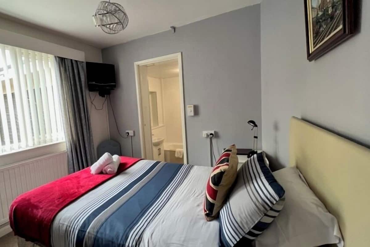 A bedroom with a bed, a TV, a window with curtains, and a door leading to a bathroom.