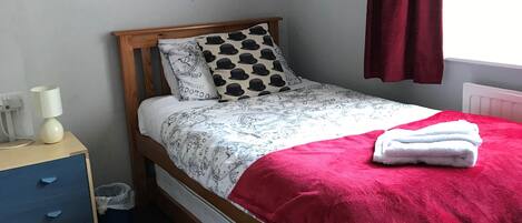 Una cama individual con estructura de madera, una colcha roja, almohadas estampadas y una mesita de noche con lámpara.