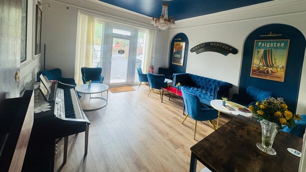 A room with a piano, blue chairs, a sofa, a table with a vase, and a poster on the wall. - The Great Western Hotel (Paignton)