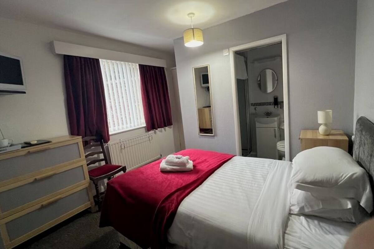 A hotel room with a bed, a dresser, a TV, a window with curtains, a mirror, and a bathroom door.
