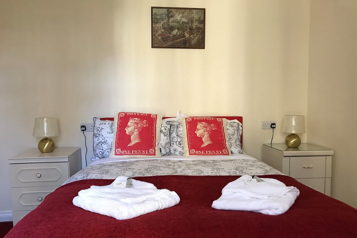 A neatly made bed with a red bedspread, two white towels, and decorative pillows with a postage stamp design. There are two bedside tables with lamps and a wall-mounted painting above the bed.