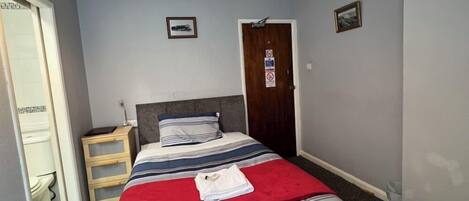 A bedroom with a bed, a wooden nightstand, a door, and a small picture on the wall.