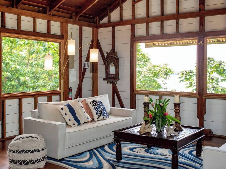 A room with a white sofa, a wooden coffee table, and a patterned rug. There are large windows with wooden frames and a view of greenery outside.