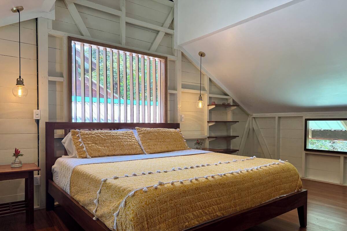 A bedroom with a wooden bed, a large window with blinds, a ceiling fan, and a TV mounted on the wall.
