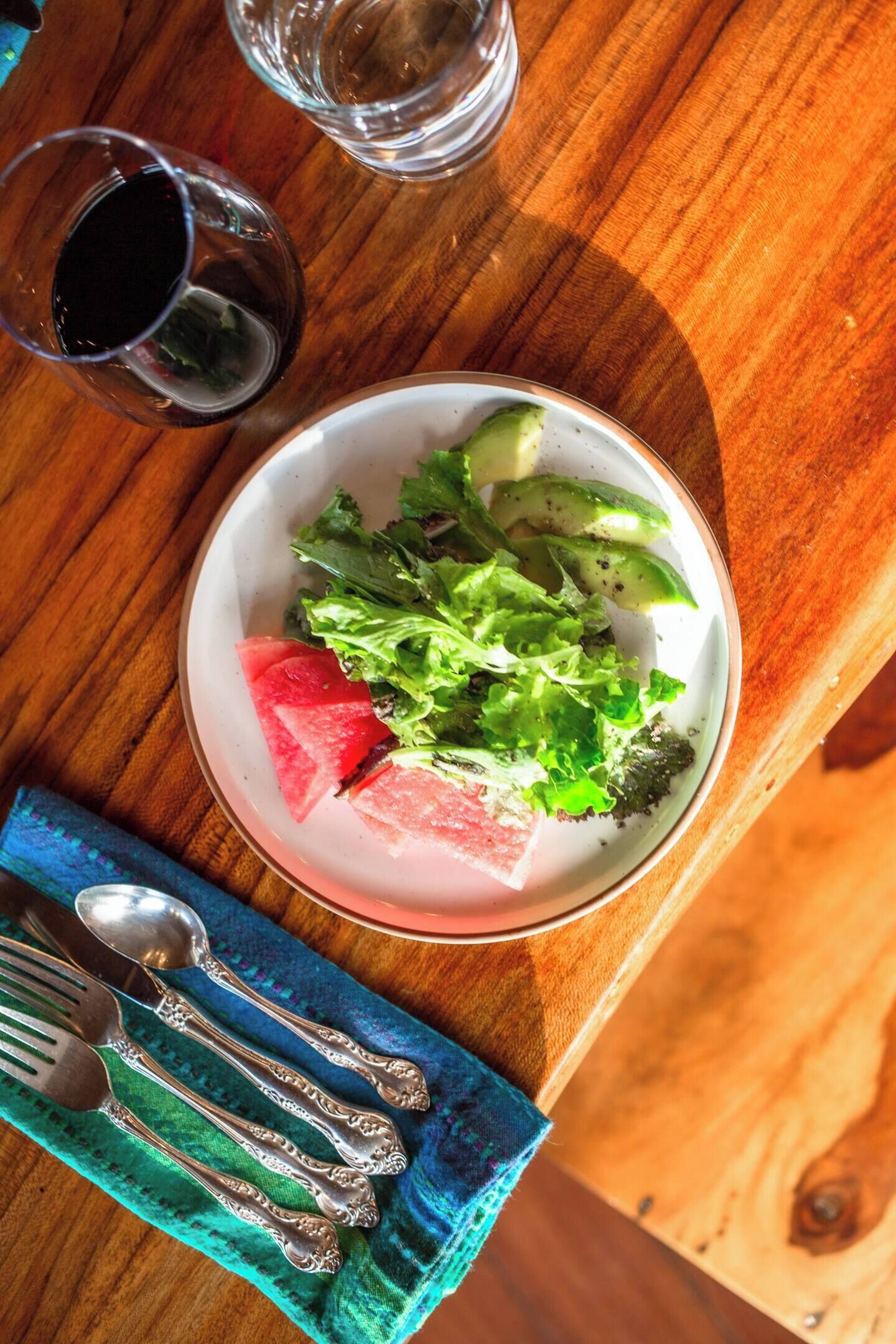 Une table en bois sur laquelle se trouve une assiette blanche contenant une salade, un verre d’eau et une cuillère, le tout posé sur une serviette bleue.