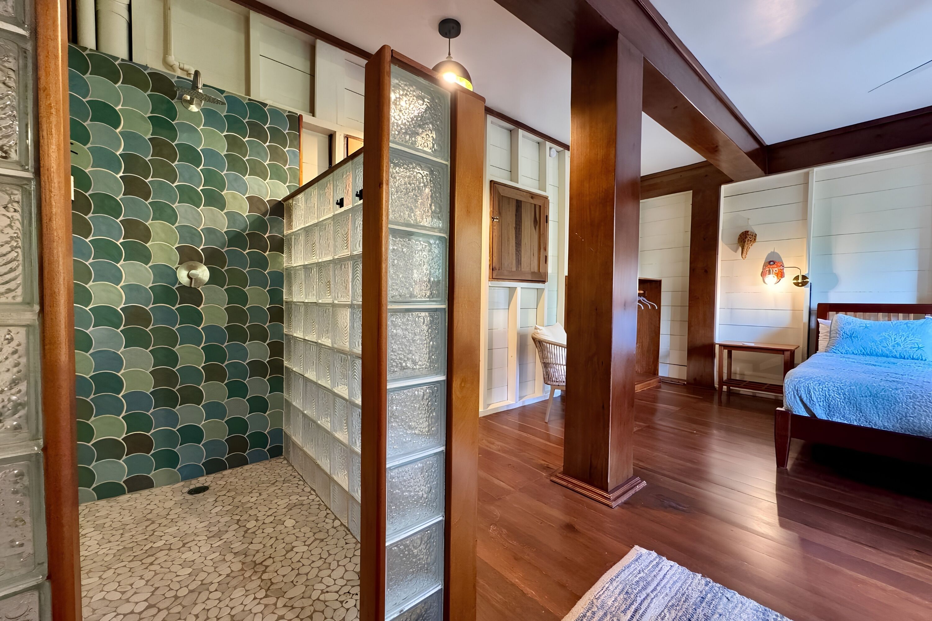 A room with a wooden floor, a bed with a blue cover, a wicker chair, and a wall with a green and white tiled pattern.