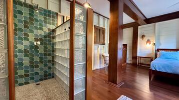 A room with a wooden floor, a bed with a blue cover, a wicker chair, and a wall with a green and white tiled pattern.