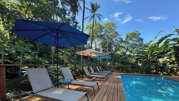 A wooden deck with lounge chairs and umbrellas, a pool, and tropical vegetation.