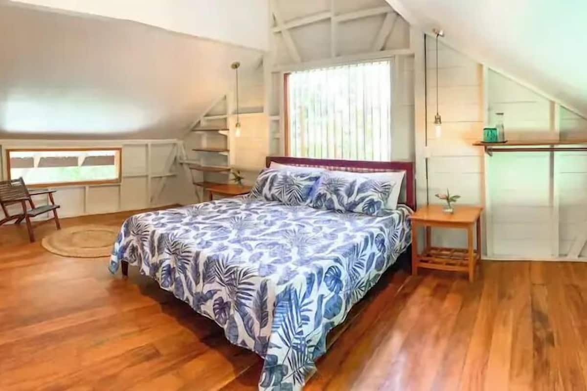 A bedroom with a bed, wooden flooring, a window, a chair, and a small table.