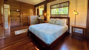 A bedroom with a wooden floor, a large bed with a blue bedspread, a window with a view of trees, and a ceiling fan. - The Lodge at Punta Rica (Bastimentos)