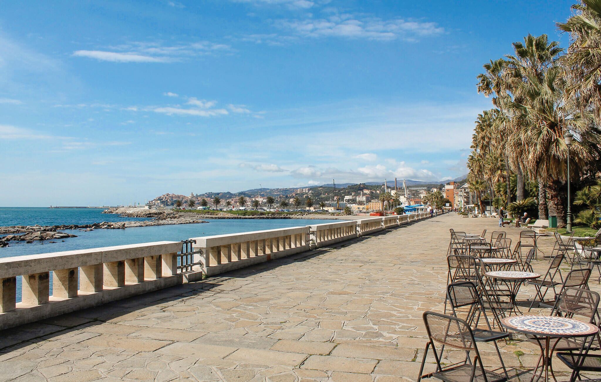 A coastal promenade with stone paving, outdoor seating, and palm trees.