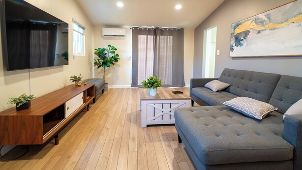 Een moderne woonkamer met een grijze bank, een houten salontafel en een flatscreen televisie aan de muur.