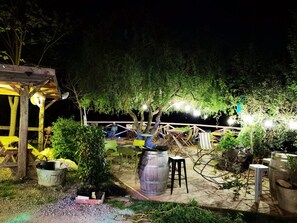 Een terras buiten met tafels en stoelen, omgeven door bomen en planten, onder een houten overkapping.