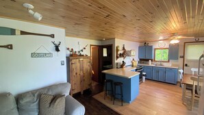 L’intérieur chaleureux d’un chalet avec un plafond en bois, une cuisine aux armoires bleues, un coin repas et un salon doté d’un canapé.