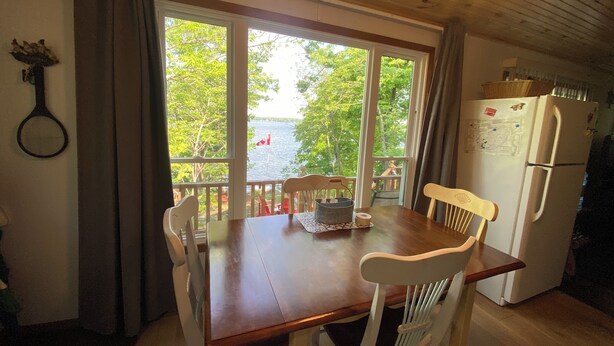 Une table à manger en bois, quatre chaises, un réfrigérateur et une vue sur un lac, visible à travers de grandes fenêtres.