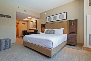 Ein Hotelzimmer mit einem großen Bett, einem Schreibtisch und einem Sessel. Über dem Bett hängt ein gerahmtes Kunstwerk, und auf dem Nachttisch steht eine Uhr.