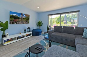 A living room with a sofa, a coffee table, a TV stand, and a large window with a view of greenery.