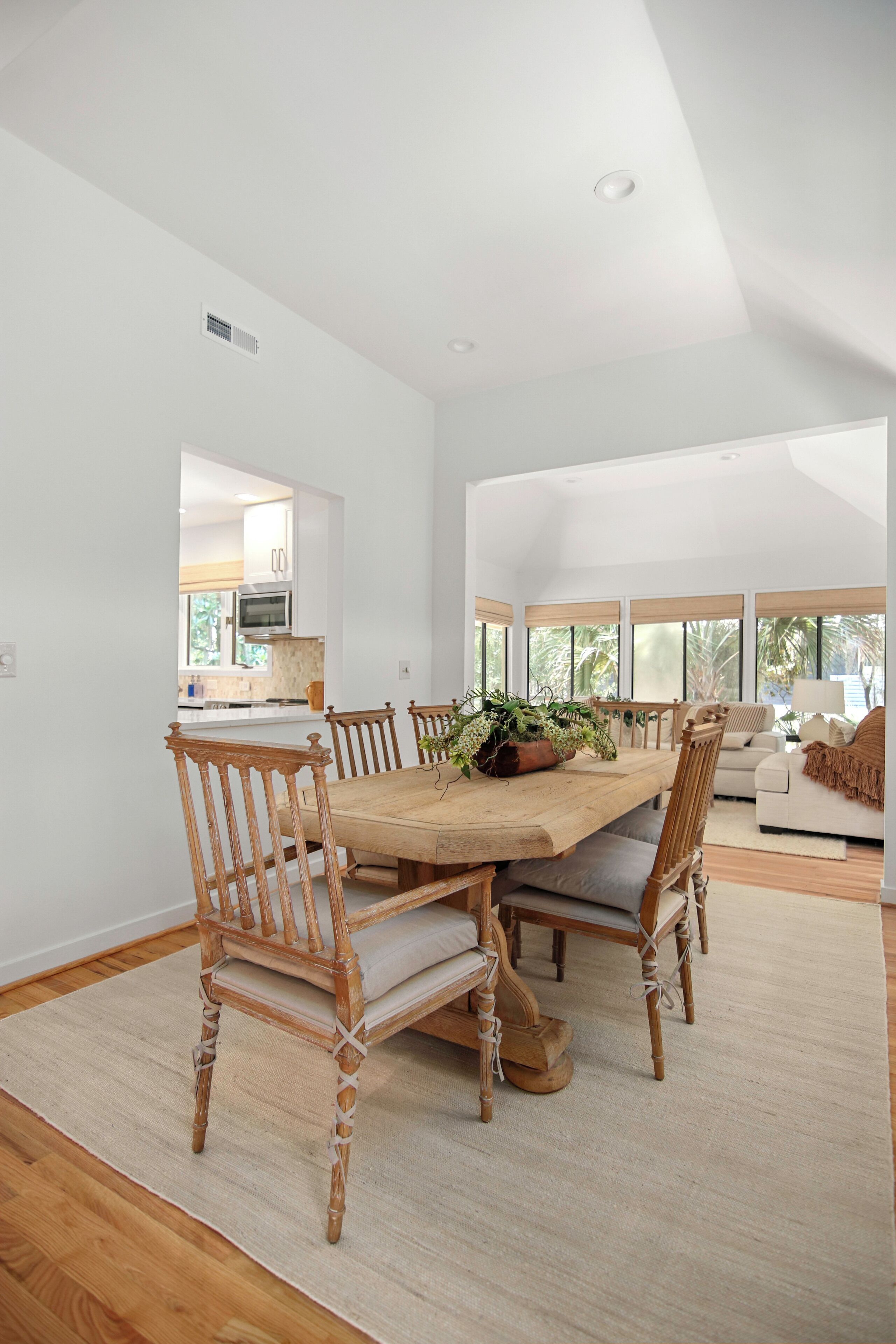 A spacious dining area with a wooden table and chairs, a large window with a view of greenery, and a kitchen in the background.