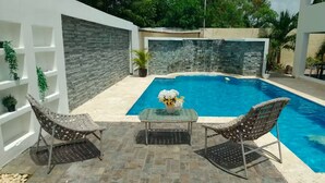 Una piscina con dos sillas de mimbre y una mesa, rodeada de muros de piedra y plantas en macetas.