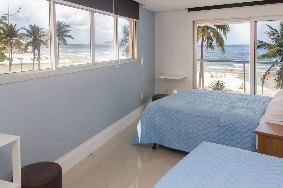 A bedroom with two beds, a window with a beach view, and a small table.