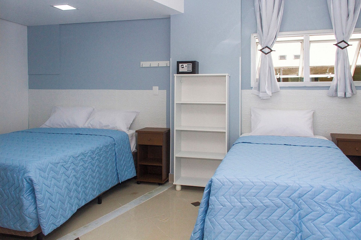 A hotel room with two beds, a white wardrobe, a small shelf unit, and a window with curtains.
