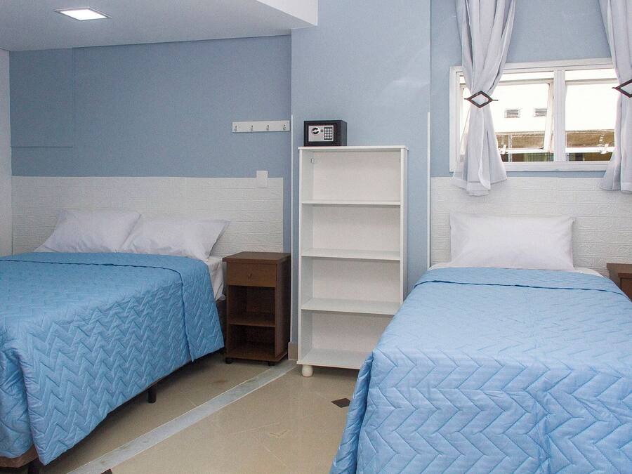 A hotel room with two beds, a white wardrobe, a small shelf unit, and a window with curtains.