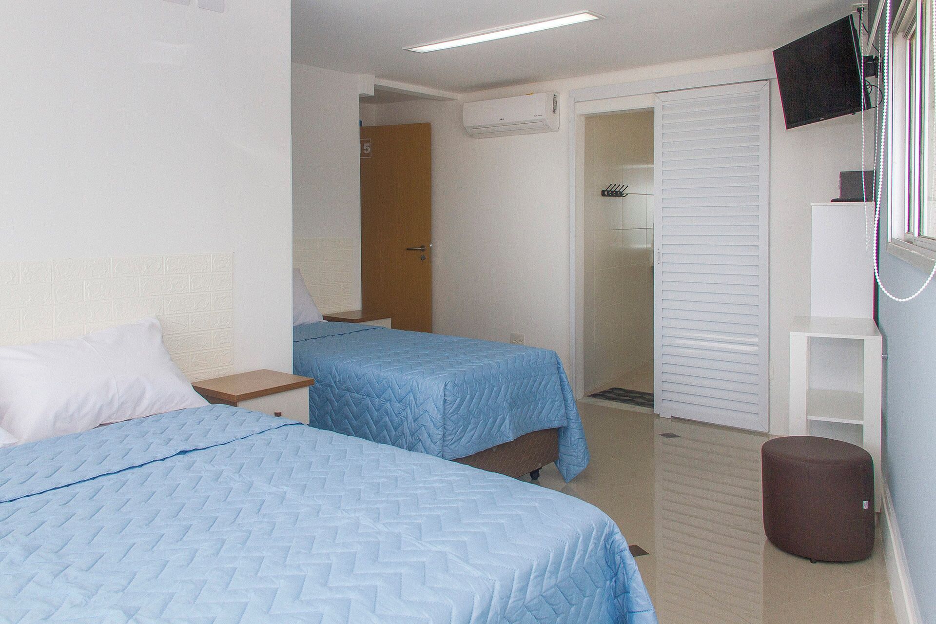 A hotel room with two beds, a TV, an air conditioning unit, and a shower area.